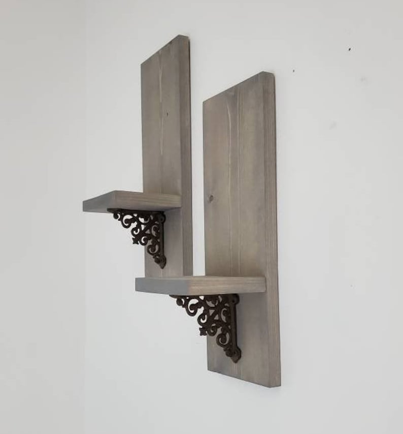 Peut inclure: Deux &eacute;tag&egrave;res flottantes en bois gris avec des supports d&eacute;coratifs en m&eacute;tal fonc&eacute;. Les &eacute;tag&egrave;res sont mont&eacute;es sur un mur blanc, mettant en valeur un design rustique. Chaque &eacute;tag&egrave;re a une forme rectangulaire et est soutenue par des supports orn&eacute;s.