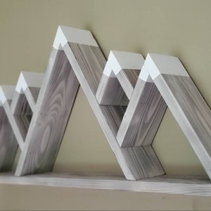 May include: A set of gray wooden shelves shaped like mountains with white painted peaks. The shelves are mounted on a wall and have a rustic, farmhouse style.