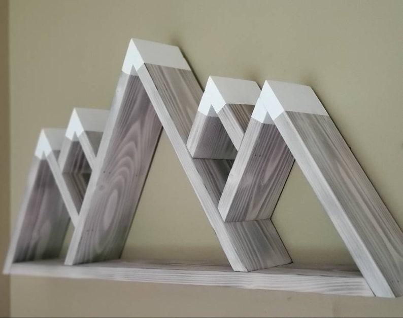 May include: A set of gray wooden shelves shaped like mountains with white painted peaks. The shelves are mounted on a wall and have a rustic, farmhouse style.
