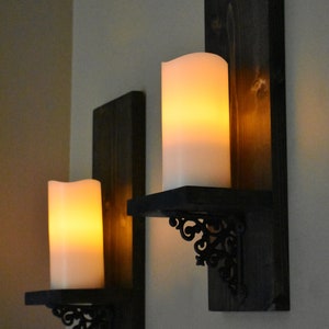 Peut inclure: Deux appliques murales de style rustique. Chaque applique comprend un fond en bois teint&eacute; fonc&eacute;, une petite &eacute;tag&egrave;re et un support d&eacute;coratif en m&eacute;tal noir. Les appliques contiennent de grandes bougies piliers blanches et lumineuses, cr&eacute;ant une ambiance chaleureuse.