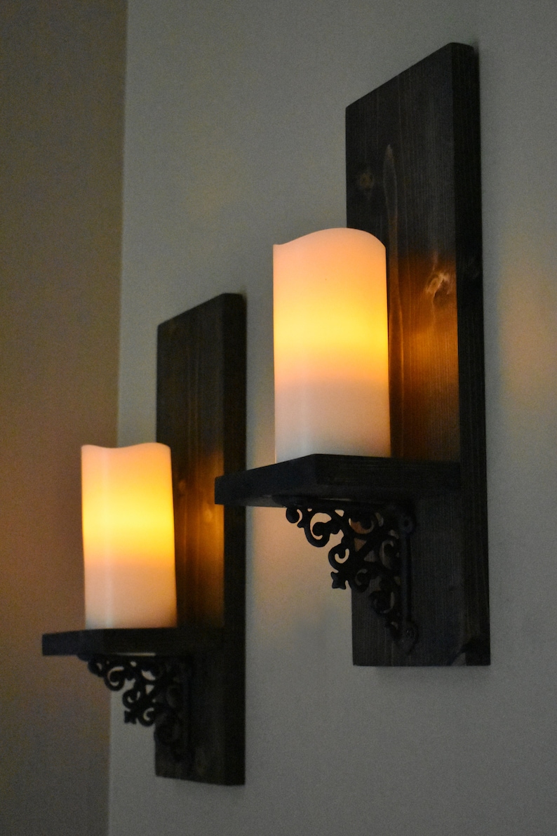 Peut inclure: Deux appliques murales de style rustique. Chaque applique comprend un fond en bois teint&eacute; fonc&eacute;, une petite &eacute;tag&egrave;re et un support d&eacute;coratif en m&eacute;tal noir. Les appliques contiennent de grandes bougies piliers blanches et lumineuses, cr&eacute;ant une ambiance chaleureuse.