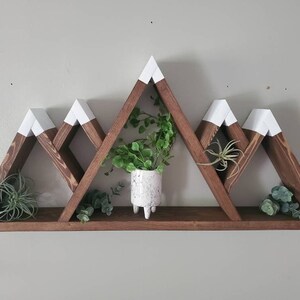 May include: A wooden mountain-shaped shelf with five triangular shelves. The shelves are painted white at the top and stained brown. The shelf is decorated with small potted plants.