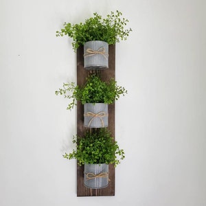 May include: A rustic wooden wall planter with three galvanized metal pots, each with a small green plant and a brown twine bow.