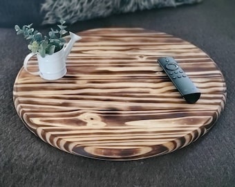 Könnte beinhalten: Eine runde Holztischplatte mit natürlicher Holzmaserung. Die Tischplatte hat ein dunkelbraunes und hellbraunes Streifenmuster. Eine kleine weiße Gießkanne mit einer grünen Pflanze steht auf der Tischplatte. Eine schwarze Fernbedienung liegt ebenfalls auf der Tischplatte.