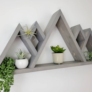 May include: Gray wooden mountain-shaped shelf with three triangular shelves. The shelf is decorated with potted plants.