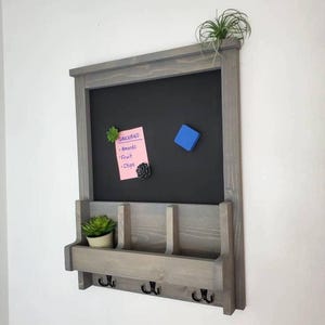 Könnte beinhalten: Eine graue Holztafel mit einem Regal, drei Haken und einem kleinen Pflanzgefäß. Ein rosa Zettel mit dem Wort "GROCERIES" und einer Artikelliste ist an der Tafel befestigt. Eine kleine Sukkulente und ein blauer Magnet befinden sich ebenfalls auf der Tafel.