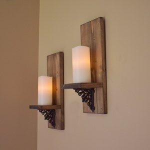 Peut inclure: Deux appliques murales rustiques en bois avec des bougies piliers blanches allum&eacute;es. Chaque applique comporte un support en m&eacute;tal sombre et d&eacute;coratif soutenant une petite &eacute;tag&egrave;re. Les panneaux arri&egrave;re en bois sont teints en brun chaud.
