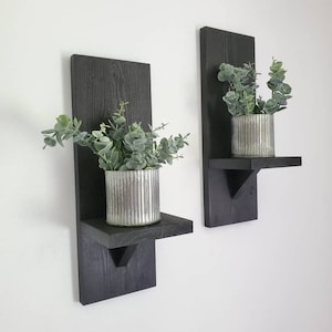 Puede incluir: Dos estantes de pared de madera negra con una peque&ntilde;a repisa y un soporte triangular. Cada estante tiene una planta en maceta con hojas verdes.