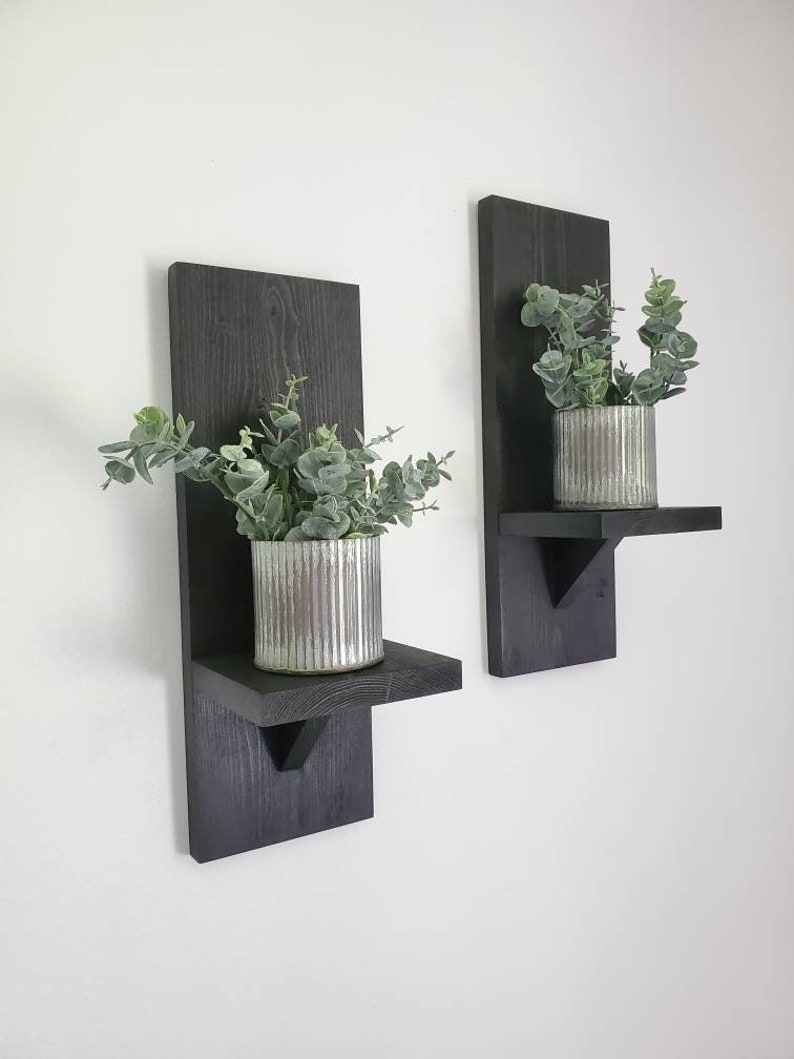 Puede incluir: Dos estantes de pared de madera negra con una peque&ntilde;a repisa y un soporte triangular. Cada estante tiene una planta en maceta con hojas verdes.