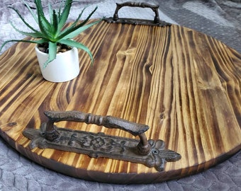 Könnte beinhalten: Ein rundes Holztablett mit zwei verzierten Metallgriffen. Das Tablett ist aus Naturholz mit einer dunkelbraunen Beize. Die Griffe sind aus schwarzem Metall mit einem dekorativen Design.