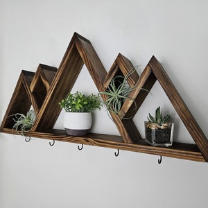 May include: A dark brown wooden mountain-shaped shelf with four triangular sections. The shelf holds small potted plants and air plants. Below the shelf are five black hooks for hanging items. The shelf is mounted on a white wall.
