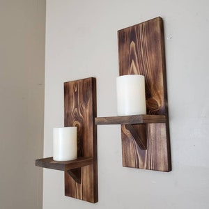 Puede incluir: Dos portavelas de madera r&uacute;stica con estantes. Cada portavelas tiene una forma rectangular y un peque&ntilde;o estante para velas. Los portavelas est&aacute;n te&ntilde;idos de un color marr&oacute;n oscuro y tienen una veta de madera natural.