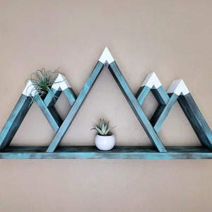 May include: Teal blue mountain-shaped wooden shelf with white peaks. The shelf has three triangular shelves and a single bottom shelf. The shelf is decorated with two small potted plants.