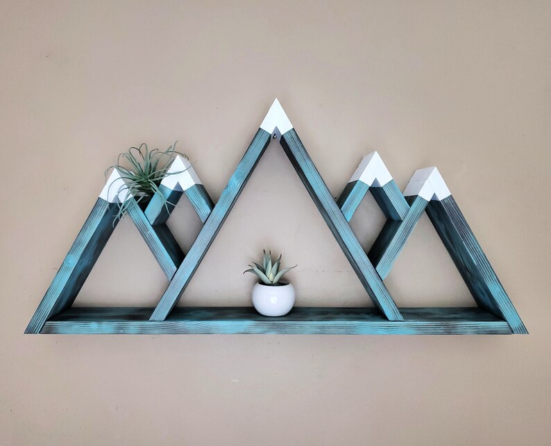May include: Teal blue mountain-shaped wooden shelf with white peaks. The shelf has three triangular shelves and a single bottom shelf. The shelf is decorated with two small potted plants.