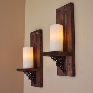 Peut inclure: Deux appliques murales rustiques avec des panneaux arri&egrave;re en bois brun fonc&eacute;. Chaque applique comprend une petite &eacute;tag&egrave;re soutenue par des supports en m&eacute;tal noir orn&eacute;, tenant une grande bougie pilier blanche. Les bougies &eacute;mettent une lueur chaleureuse.