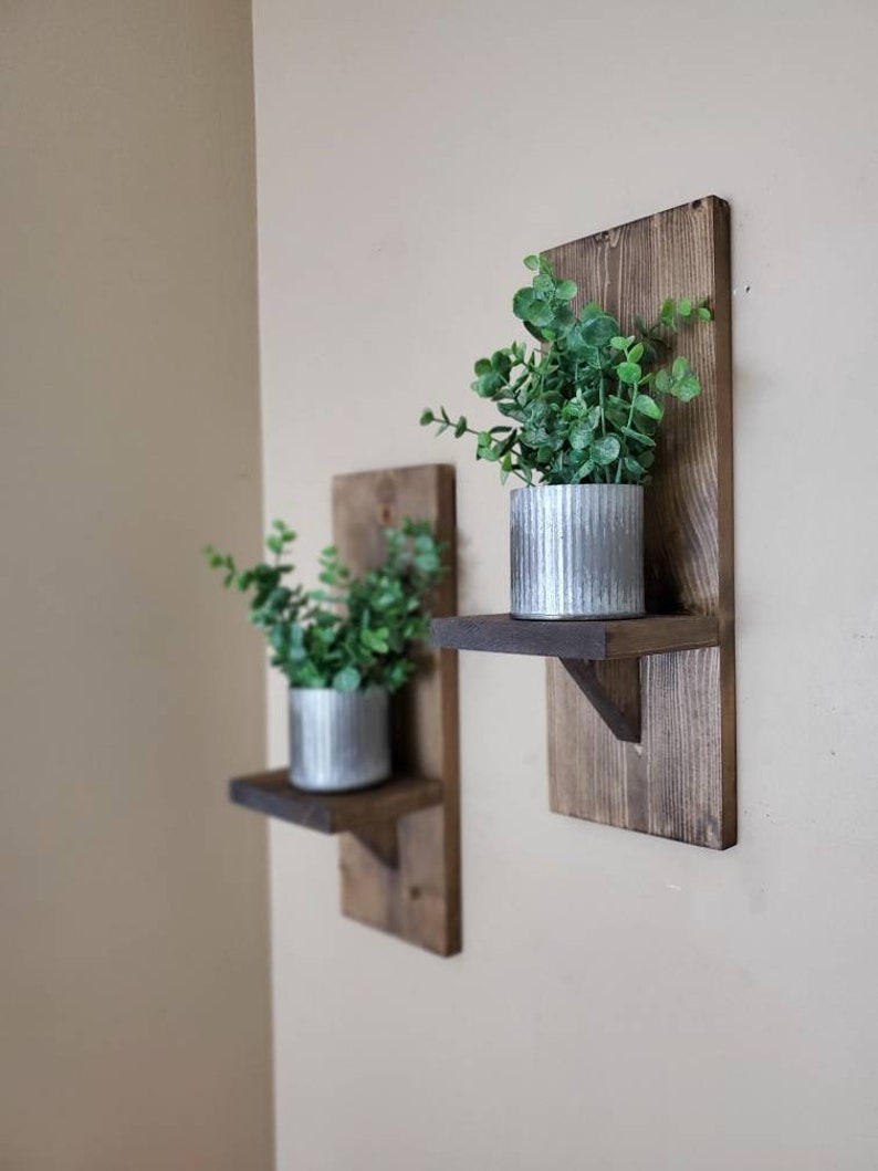 Puede incluir: Dos estantes de pared de madera r&uacute;stica con una peque&ntilde;a repisa, cada uno con una planta en maceta con hojas verdes en una maceta de metal plateado. Los estantes est&aacute;n te&ntilde;idos de un color marr&oacute;n oscuro.