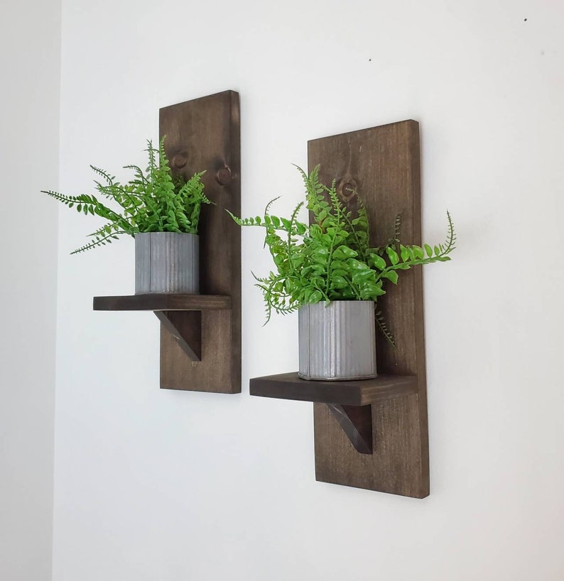 Puede incluir: Dos estantes de pared de madera marr&oacute;n oscuro con una peque&ntilde;a repisa y una maceta de metal en cada estante. Los estantes est&aacute;n montados en una pared blanca y cada estante tiene una planta verde en la maceta.