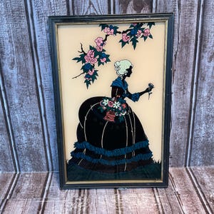 May include: Framed silhouette art featuring a woman in a period dress holding a flower. The black silhouette is set against a cream background, with pink and blue floral accents. The frame is black and the artwork is displayed on a wooden surface.