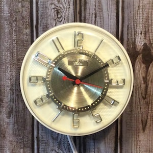 Puede incluir: Un reloj de pared vintage con una esfera blanca y un centro plateado. El reloj presenta manecillas negras, un segundero rojo y marcadores de hora plateados. El reloj está encerrado en un marco blanco y cuelga sobre un fondo de madera.