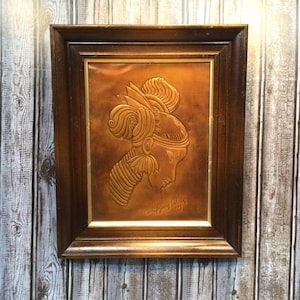 May include: A framed copper relief art piece featuring a profile of a woman with an elaborate headdress. The artwork is in a dark brown wooden frame with a gold inner border. The relief is a warm copper color.