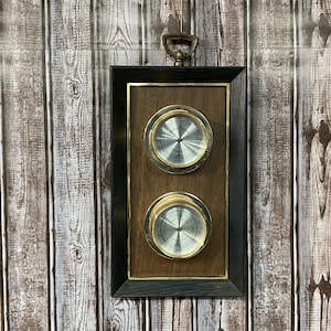 Puede incluir: Un barómetro rectangular con marco de madera oscura y dos medidores circulares. Los medidores tienen caras plateadas y bordes dorados. El fondo es marrón. Un gancho de color latón en la parte superior para colgar. Mide unos 30 cm de alto.
