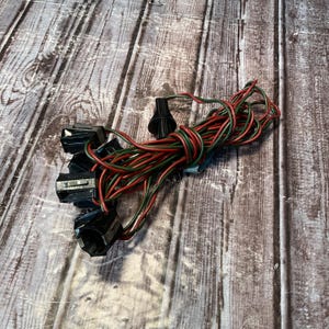 May include: A set of black plastic light sockets connected by a series of intertwined red and green electrical wires. The assembly includes a single black plastic conical plug at the end of the wire bundle, designed for electrical connectivity.