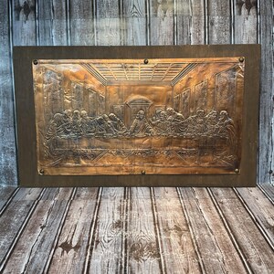 May include: A rectangular copper wall plaque featuring a relief depiction of The Last Supper. The metallic artwork is mounted on a dark brown wooden board and secured with six small metal nails along the edges of the copper panel.