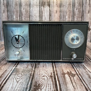 Puede incluir: Una radio reloj General Electric vintage. La esfera del reloj está a la izquierda, con las palabras "SOLID STATE" encima. El dial de la radio está a la derecha, con un mando de volumen debajo. La radio es negra y plateada.