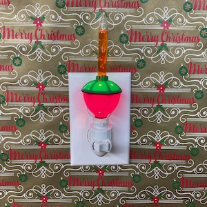 May include: A plug-in night light featuring a white base, a bright pink rounded bulb, and a clear orange bubble light tube on top. The device is mounted on a white rectangular wall plate, creating a festive holiday lighting display.