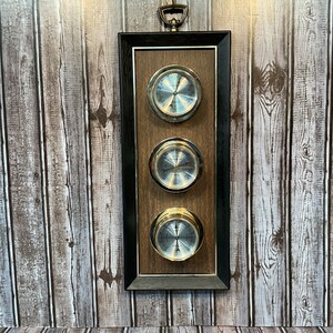 Puede incluir: Una estación meteorológica rectangular de madera montada en la pared con tres medidores circulares. Los medidores tienen bordes y caras de metal plateado y dorado. El marco es negro con un respaldo de madera marrón. Un asa decorativa de metal en la parte superior.