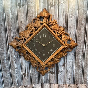 May include: A decorative wall clock with a square face and a brown wooden frame. The frame is adorned with carved grape vines and leaves. The clock face is black with gold numerals and hands. The clock is mounted on a wooden wall.