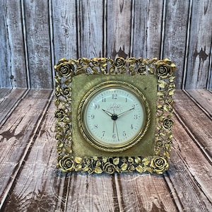 May include: A square analog clock featuring a gold-toned frame decorated with intricate sculpted roses and leaves. The clock face is off-white with black numerals and a central floral illustration, surrounded by a textured gold-toned square border.