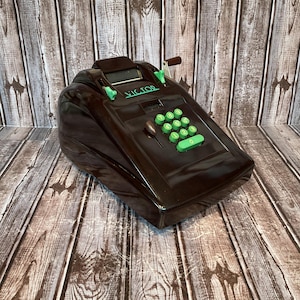 Op de afbeelding: Een vintage bruine "Victor" optelmachine met groene toetsen en accenten. De machine heeft een slinger en een klein displayvenster. Het woord "Victor" staat in het groen op de voorkant. Op een houten oppervlak.