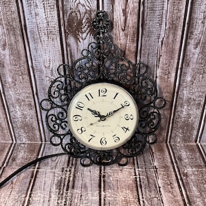 May include: Black, ornate wall clock with a cream-colored face and black hands and numbers. The clock is surrounded by a decorative, scrollwork frame. The clock face reads "General Electric".