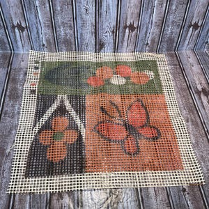 May include: A needlepoint canvas with a grid pattern, featuring four distinct sections. The top left section is green, the top right has red shapes, the bottom left has an orange flower, and the bottom right has an orange butterfly.