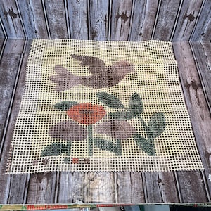 May include: Needlepoint canvas featuring a floral and bird design. The design includes a red flower, green leaves, and a brown bird against a cream-colored background. The canvas is made of a grid-like material, ready for stitching.