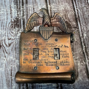 Puede incluir: Una cubierta de interruptor de luz de tono cobrizo vintage con un emblema de águila y un pergamino con una inscripción. La placa del interruptor tiene dos interruptores de palanca y está montada sobre una superficie de madera. El texto del pergamino dice "PEACE".