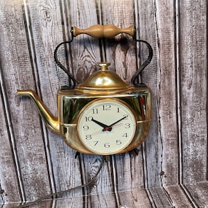 May include: A gold-colored metal wall clock shaped like a tea kettle. The clock face is cream-colored with black numerals, black hour and minute hands, and a red second hand. It features a black metal handle with a light brown wooden grip.