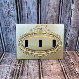 Puede incluir: Una placa de interruptor de luz dorada y ornamentada con tres aberturas rectangulares. La placa presenta un diseño ovalado con adornos decorativos y un águila en la parte superior. El fondo es una superficie de madera.