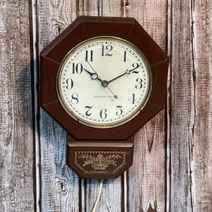 Op de afbeelding: Een vintage bruine achthoekige wandklok met een crèmekleurige wijzerplaat en goudkleurige rand. De wijzerplaat toont zwarte cijfers en wijzers, met de tekst "GENERAL ELECTRIC" eronder. Een decoratief onderste gedeelte heeft een bloemenontwerp.