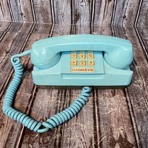 May include: A light blue Starlite telephone with a matching coiled cord. The device features a rectangular base, a handset resting on top, and a central keypad with beige buttons displaying numbers, letters, and symbols. The brand name Starlite is printed in black text below the keypad.