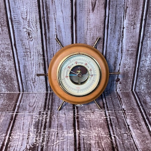 May include: A circular wooden barometer featuring a brass-toned rim and six protruding metal spokes resembling a ship's wheel. The white dial displays the text Stormy, Rain, Change, Fair, and Very Dry, with a central needle and a small brass gear.