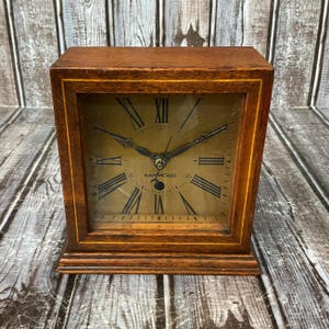Op de afbeelding: Een vintage houten klok met een vierkante wijzerplaat en Romeinse cijfers. De wijzerplaat is lichtgoudkleurig en de wijzers zijn zwart. De klok is gemarkeerd met "HAMMOND" en heeft een donkerbruin houten frame.