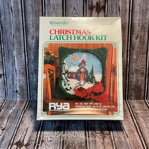 May include: A WonderArt Christmas Latch Hook Kit in a white box. The box features a graphic of a finished square pillow with a winter scene, a house, and red poinsettias. Text includes 6 1/2 oz. net weight and a finished size of 15x15 inches.