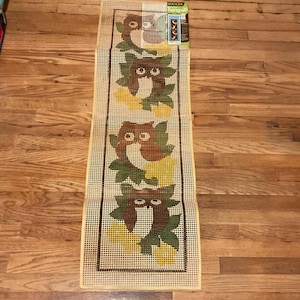 Op de afbeelding: Een rechthoekig borduurcanvas met een herhalend patroon van bruine uilen, groene bladeren en gele accenten. De uilen staan tegen een beige achtergrond met een zwarte rand. De verpakking bevat een "BUCILLA hang-up" label.