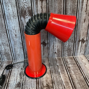 Puede incluir: Una lámpara de escritorio roja y negra con una base cilíndrica y un cuello flexible y acanalado. La lámpara tiene una base redonda y una pantalla roja. La lámpara está enchufada a un cable de alimentación negro.