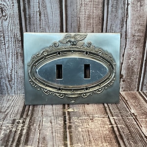 Puede incluir: Una placa de interruptor de luz de estilo vintage plateado con un centro ovalado y dos aberturas rectangulares. La placa presenta un borde ornamental en relieve con un águila en la parte superior. El fondo es una superficie de madera.