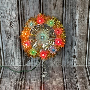 May include: A circular Christmas tree topper featuring a metallic silver starburst centre surrounded by a ring of gold tinsel. The ornament is decorated with small, star-shaped light covers in red, green, and blue, mounted on a silver metal coil base.