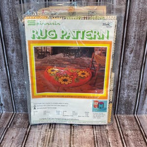 Könnte beinhalten: Ein Vintage-Teppichmuster-Kit in einer Plastikverpackung. Das Kit zeigt ein farbenfrohes Blumendesign mit rotem Hintergrund und gelben und grünen Blumen. Die Verpackung enthält den Text "Spinnerin Rug Pattern". Die Abmessungen betragen 63,5 x 101,6 cm.