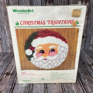 May include: A WonderArt Christmas Traditions latch hook wall hanging kit featuring a round Santa Claus design. The packaging displays the finished 27-inch round project, including pre-cut acrylic yarn, a printed canvas, and an illustrated guide.